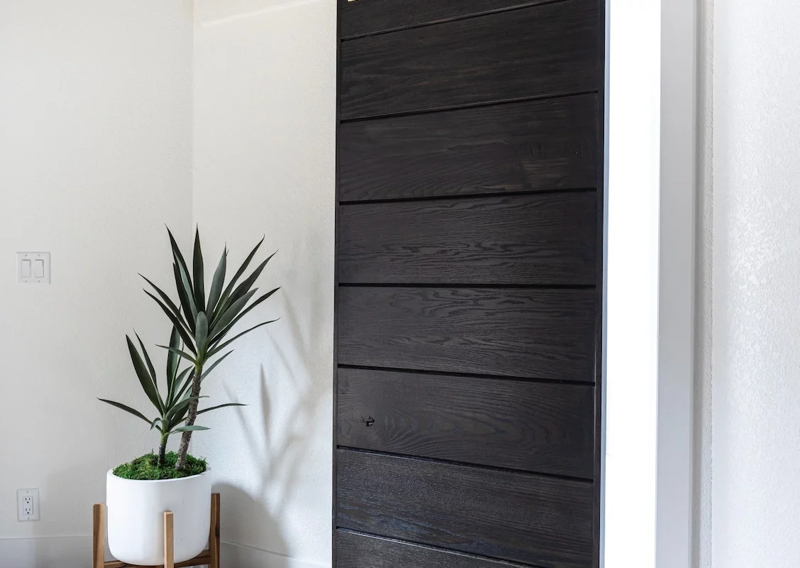 A dark wooden sliding barn door installed by a general contractor in Bend, Oregon during a bathroom remodel. The door is on a golden rail and partially open, situated beside a potted plant in the modernly styled room with light wooden flooring.